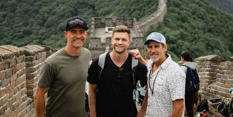 Three men stand together on the Great Wall of China during their visit to China 2024. The man on the left wears a green shirt and red shorts, the middle man wears a black shirt and shorts, and the right man dons a patterned shirt and shorts.